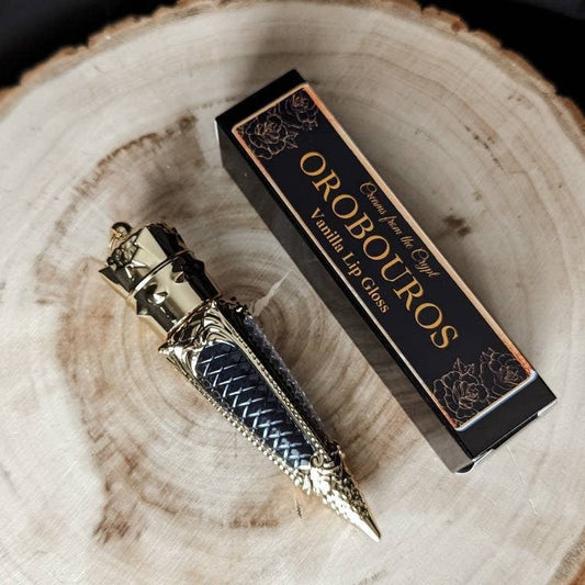 A luxurious gold bottle of OROBOUROS black lip gloss with a French vanilla design, displayed next to its packaging on a wooden surface.