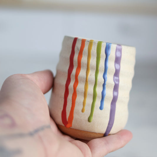 "Hands cupping cream colored ceramic wine cup featuring rainbow drip glaze pattern with red, orange, yellow, green, blue and purple flowing down the sides"