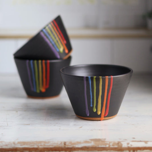 "Three black ceramic bowls stacked at angles showing rainbow drip glaze patterns with vibrant colored stripes against dark background"