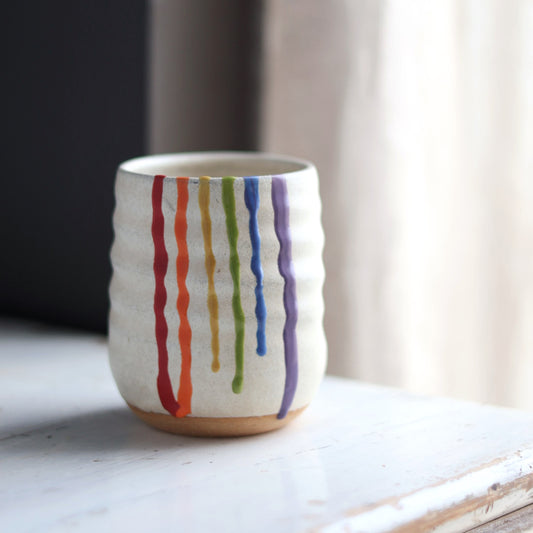 "Cream colored ceramic wine cup featuring rainbow drip glaze pattern with red, orange, yellow, green, blue and purple flowing down the sides"