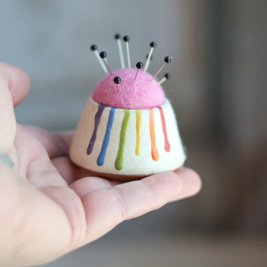 "Ceramic pin cushion with rainbow drip glaze topped with pink felted wool and multiple sewing pins, held in palm of hand"