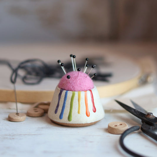"Ceramic pin cushion with rainbow drip glaze topped with pink felted wool and multiple sewing pins, held in palm of hand"