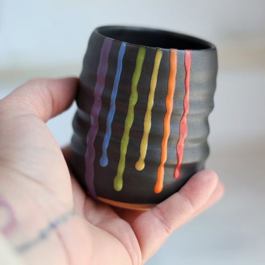 "Black ceramic wine cup with textured surface and rainbow drip glaze held in a hand, showing vibrant colored stripes"