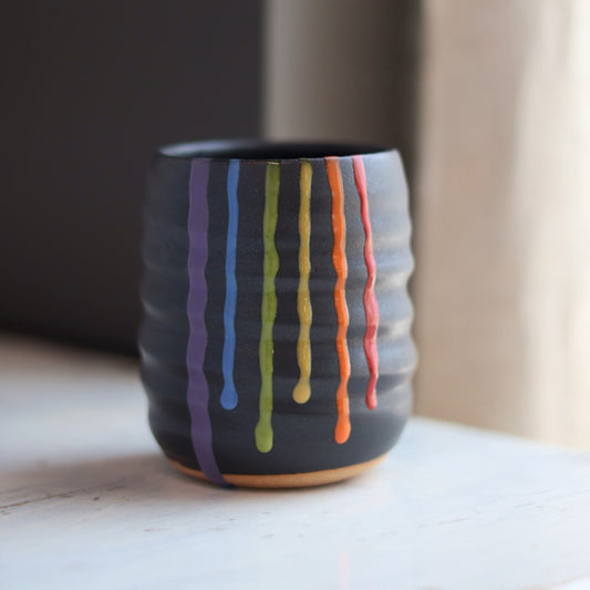 "Black ceramic wine cup with textured surface and rainbow drip glaze showing vibrant colored stripes"